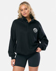 A woman wearing a black snap-collar varsity sweatshirt with a chest logo, paired with black shorts.