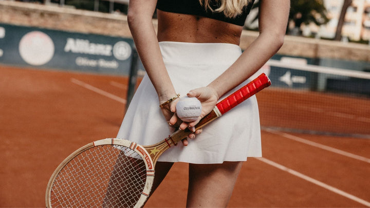 Woman Wearing White Tennis Skort and Black Sports Bra, Facing away from camera holding a tennis ball and tennis racket